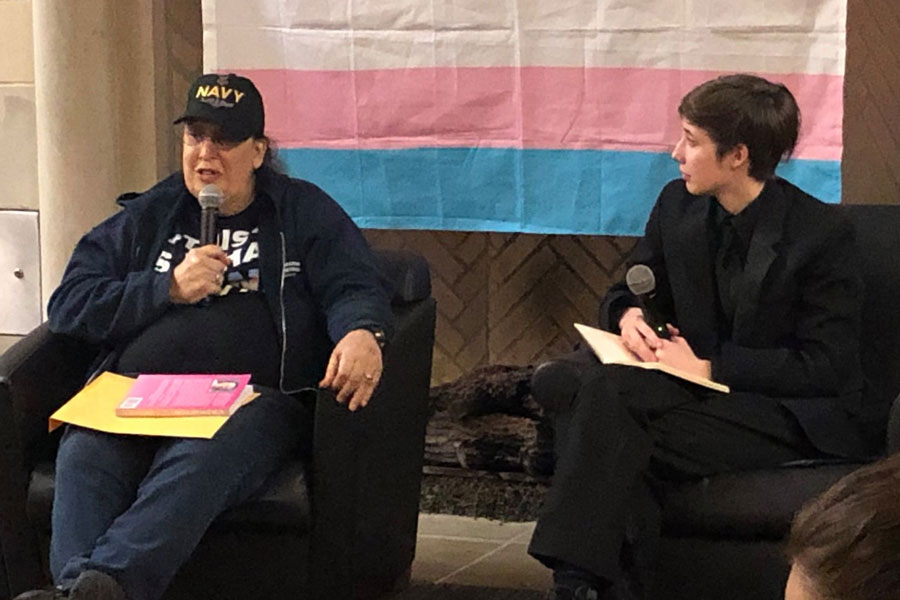 Two people sit in front of the transgender pride flag and address an audience.