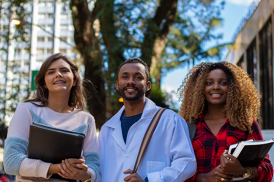 Three students in a row smiling, two students holding textbooks, and one student holding backpack strap.