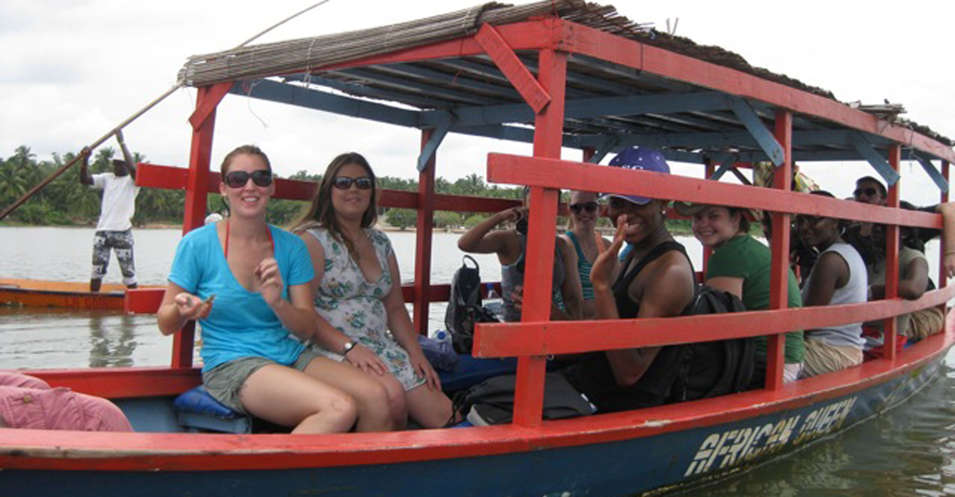 Students sit in a boat on the water and wave at the camera.