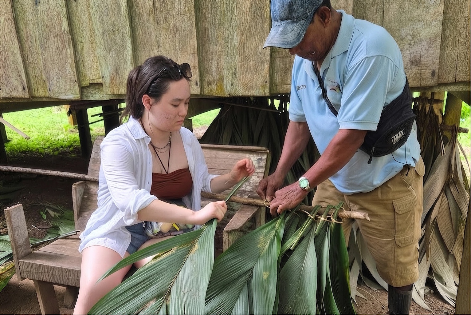 An Agnes Scott students working with a man to bind large leaves to a wooden stick for a roof. 