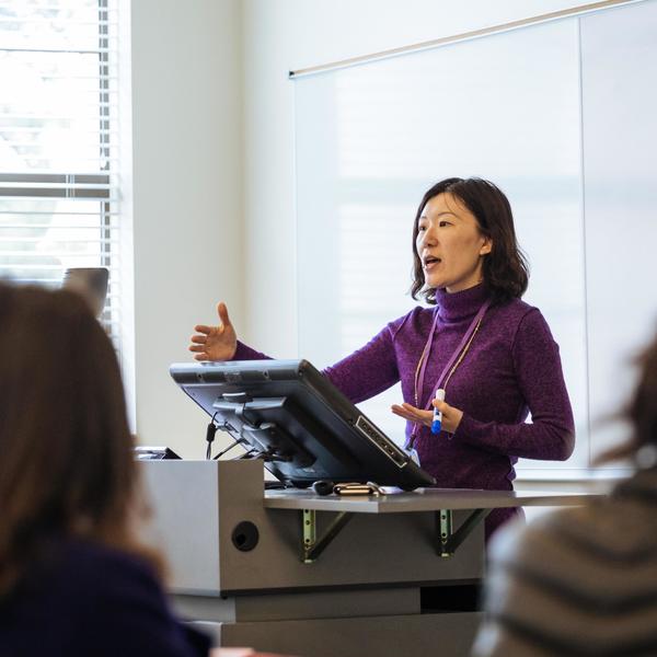 An economics major professor lectures her class at a podium.
