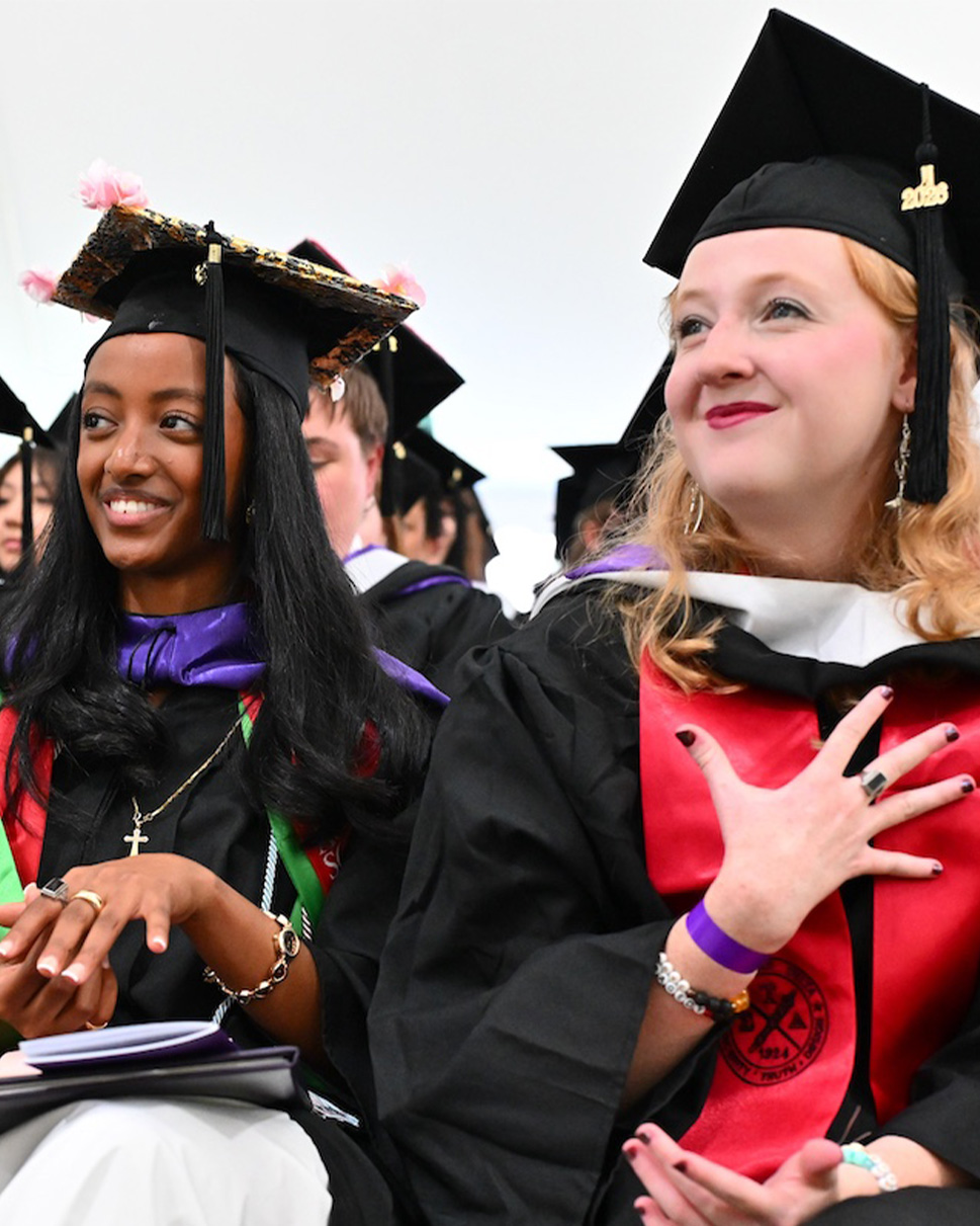 two Agnes Scott students sitting at graduation and showing their rings