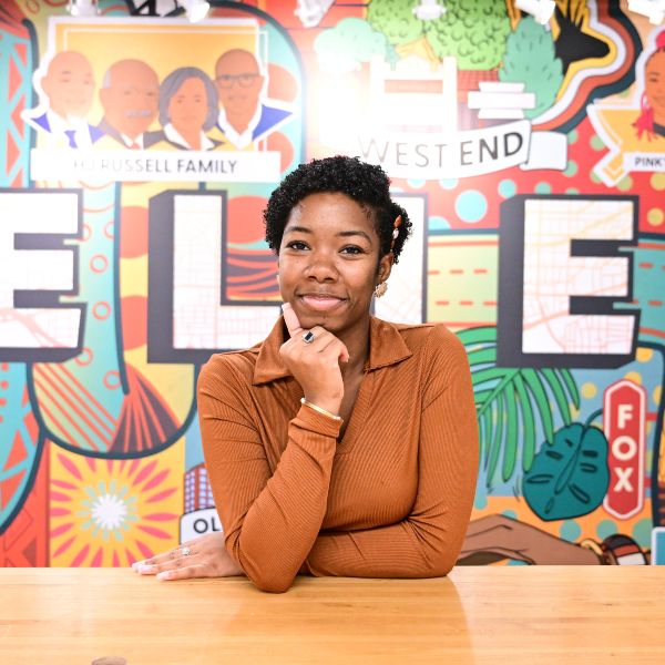 A confident Agnes Scott College student sits at a table in front of a colorful mural in Atlanta’s West End, smiling with her hand resting under her chin.