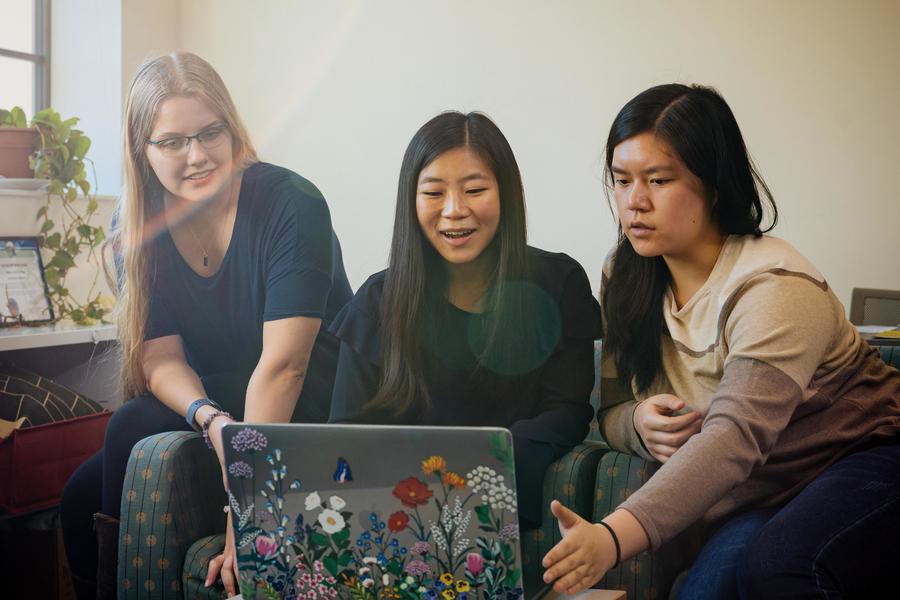 Three students look at a laptop while sitting on a bed.