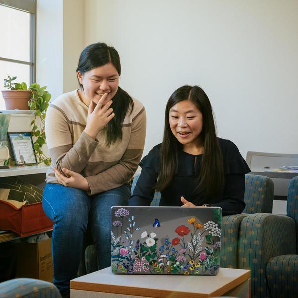 Two students look at a laptop with stickers of flowers and laugh at what they see.