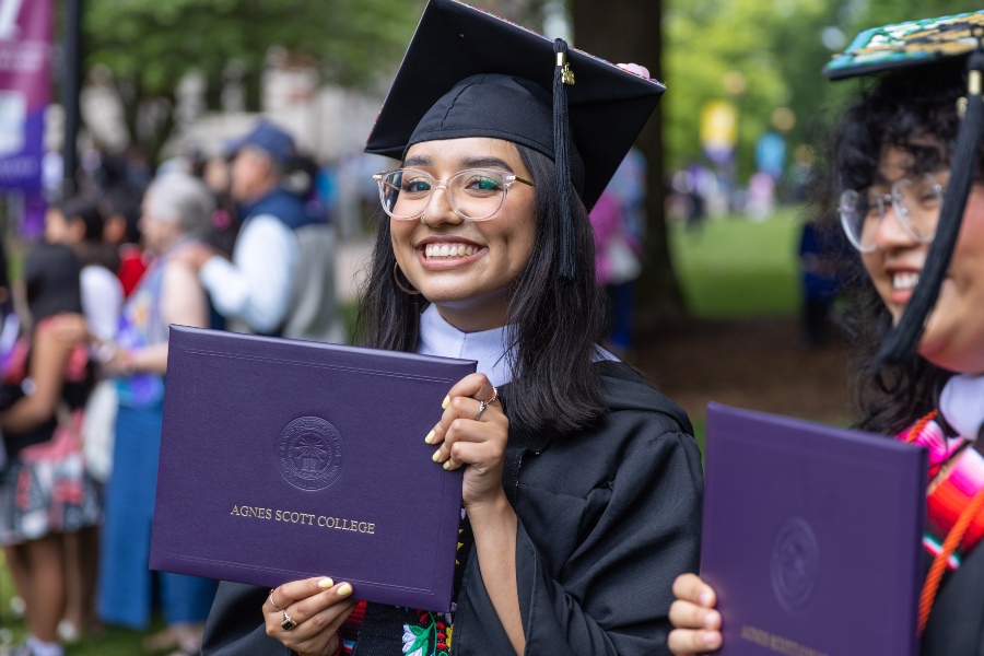 A graduate with glasses holds their diploma proudly while looking at the camera