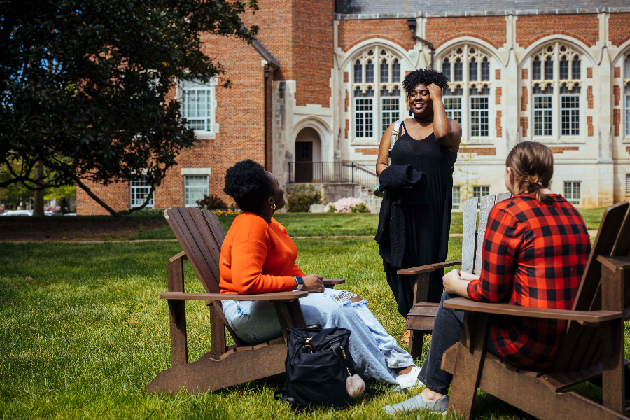 students sit in wooden chairs, two with their back turned to the camera as a woman speaks to them