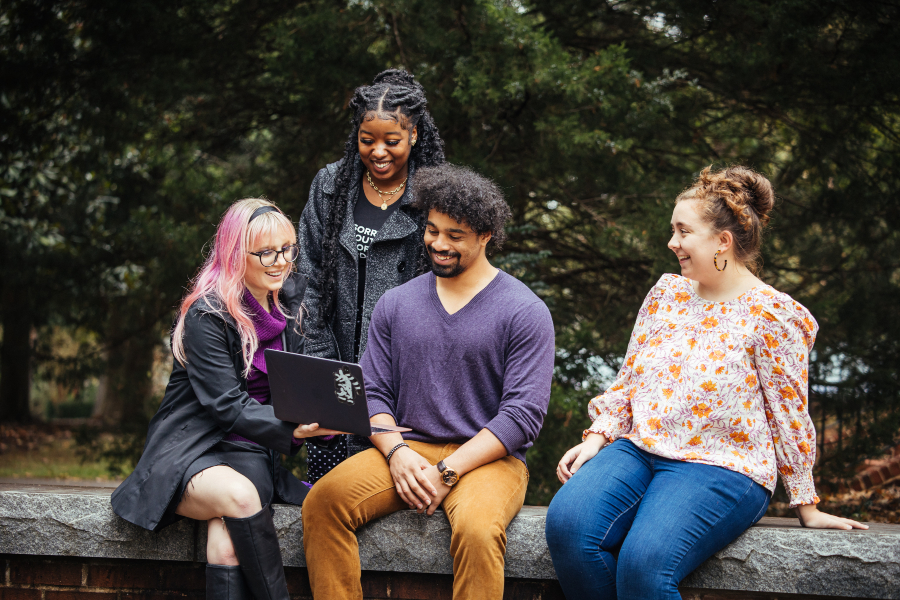 group of students sit on a ledge in fall clothing while looking at a laptop screen