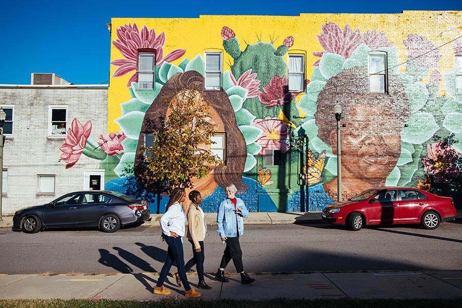 group of three students walking down sidewalk in front of bright mural of two faces