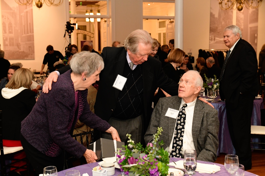 Three people welcome each other at a table with a purple tablecloth covering it.