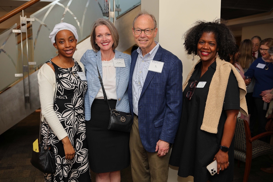 Four attendees at Agnes Scott College’s scholarship donor event pose together and smile for the camera. The group includes a student and three guests wearing nametags, standing in front of a staircase inside a warmly lit venue filled with other attendees in the background.