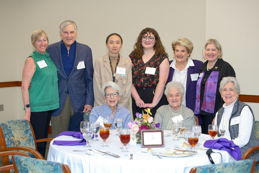 A group of scholarship donors, alumnae, and Agnes Scott students gather around a table marked with a “5” at a donor recognition event. Four people are seated at the table with glasses of iced tea and floral centerpieces, while six others stand behind them, all smiling warmly for the photo in a brightly lit event space.