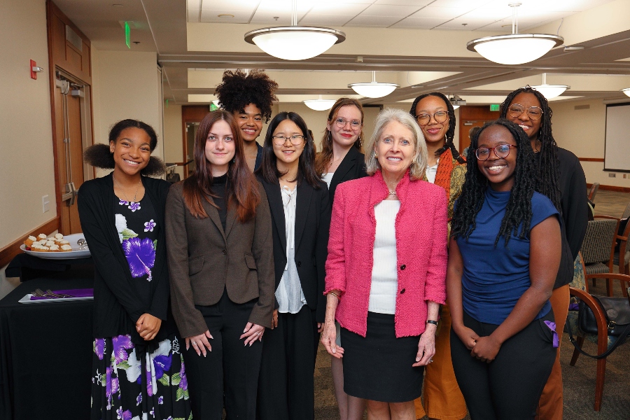 A group of Agnes Scott College students and a donor pose together at a scholarship recognition event. The group includes nine women, smiling and dressed in a mix of professional and semi-casual attire, standing in a warmly lit event space with round tables and overhead lighting in the background.