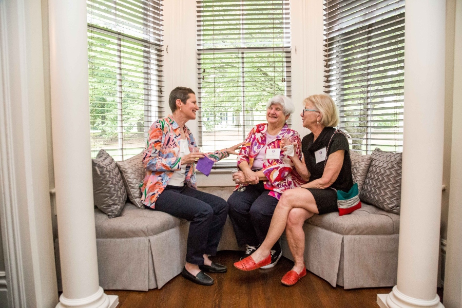 Three people talk while holding drinks and sitting on a sectional couch inset of a window well. 