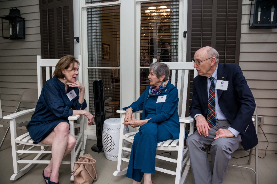 Three people sit on porch rocking chairs while talking. 