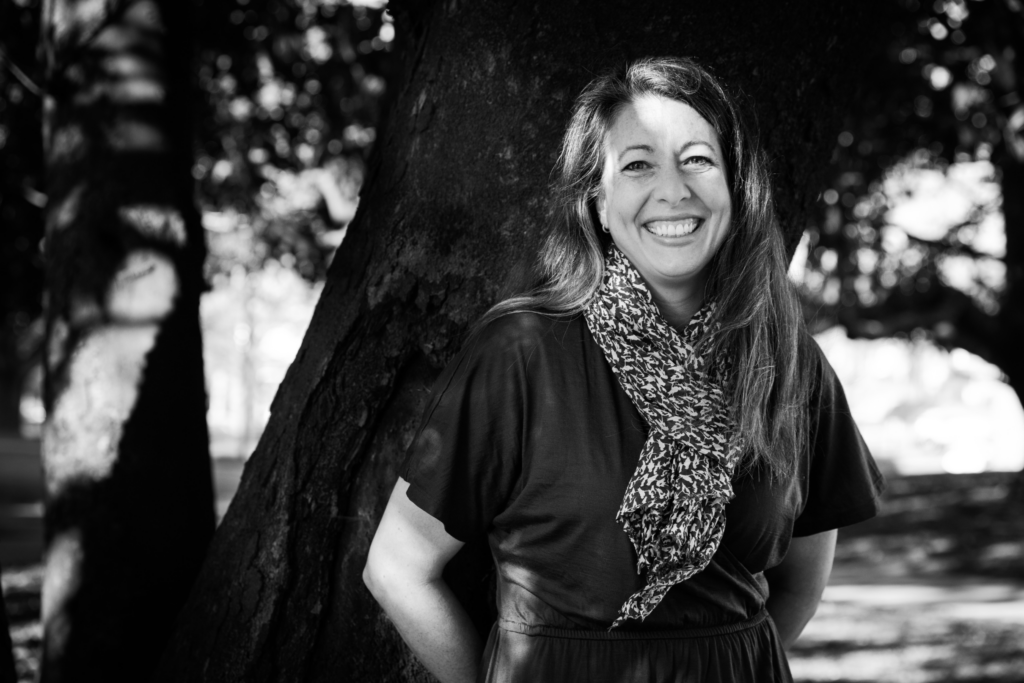 A black and white portrait of a smiling woman with long, wavy hair, standing outdoors leaning against a large tree trunk. She is wearing a dark short-sleeved dress and a patterned scarf. Sunlight filters through the trees, casting dappled shadows on the ground and the tree bark. The background consists of additional trees and blurred foliage.